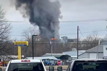 Thick black smoke rises from a factory fire in the distance, viewed from a roadway with vehicle tops in the foreground.