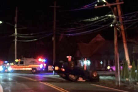Overturned car on a dark road at night, with emergency vehicle lights flashing in the background.