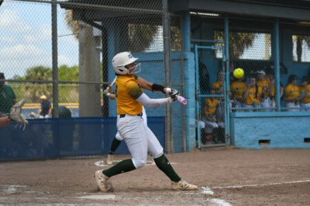 Softball player in yellow and green uniform swings a pink bat at a yellow pitch on a dirt field.