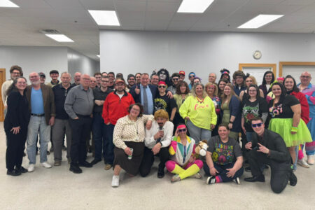 A large group of people, some in 80s-themed costumes, pose for a photo at an event.