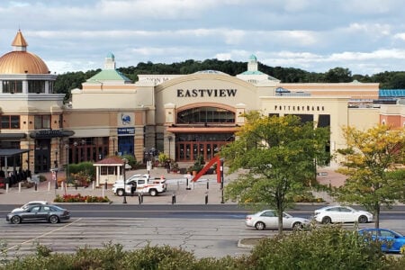 Exterior view of Eastview Mall in Victor, NY, showing its main entrance, parking lot, and surrounding trees under a cloudy sky.
