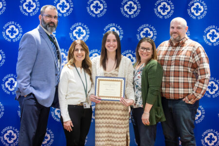 A smiling student holds an award certificate, flanked by four adults, in front of a Finger Lakes-Wayne BOCES backdrop.