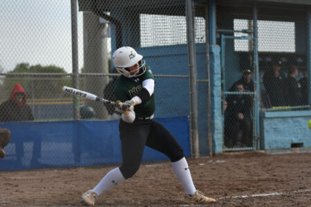 A Keuka College softball player in a green and white uniform swings a bat during a game.