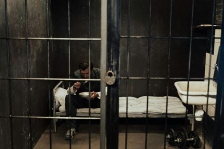 Man eats from a bowl on a bed in a dark jail cell behind metal bars.