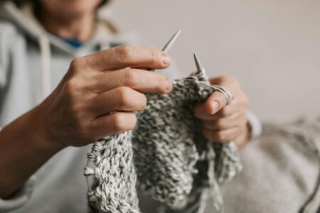 Person in gray hoodie knits gray and white yarn with silver needles indoors.
