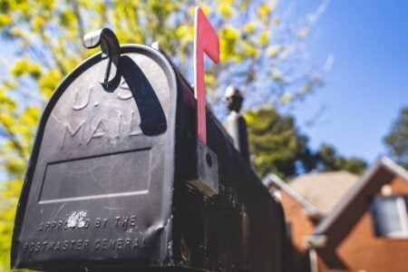 A black U.S. Mail mailbox with its red flag raised, against a blue sky and blurred green foliage.