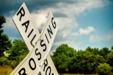 A white railroad crossing sign with black text against a blue sky with clouds and green trees in the background.
