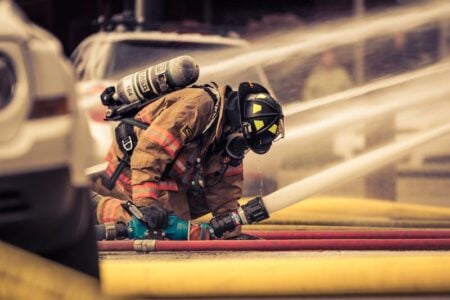 Firefighter in full gear kneels, connecting hoses with water streams and other hoses visible in the background.