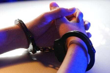 Hands in handcuffs rest on a surface, illuminated by blue and white light.