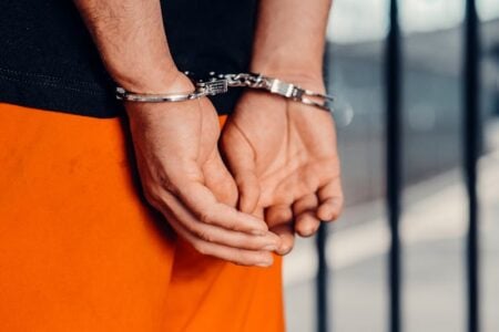 Hands of a person in orange jail uniform, cuffed behind their back, with blurred prison bars in background.