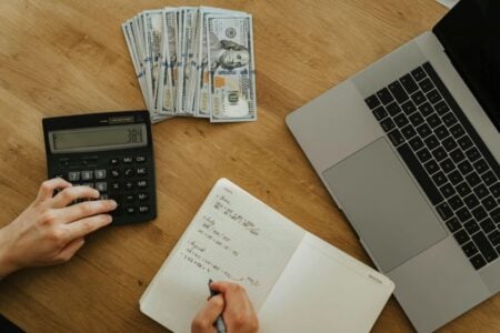 Hands use a calculator and write in a notebook, with a laptop and stacks of hundred-dollar bills on a wooden desk.
