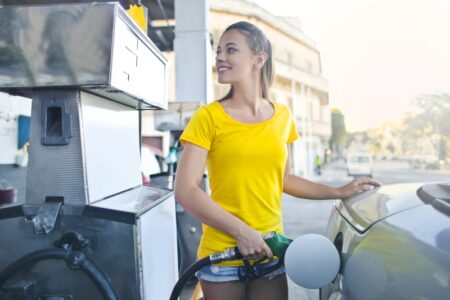 Smiling woman in a yellow shirt pumps gas into her car at an outdoor gas station.