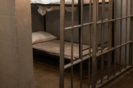 Bunk beds are seen through the metal bars of a jail cell, with white bedding.