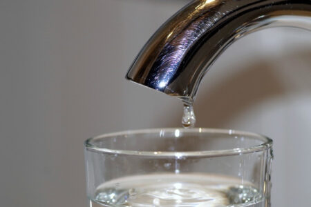 A drop of water falls from a chrome faucet into a glass.