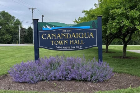 Canandaigua Town Hall sign at 5440 Routes 5 & 20W features gold lettering and a lake graphic with sailboats above flowering plants.