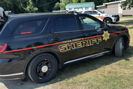 A black Yates County Sheriff SUV with gold lettering parked on grass, with another sheriff vehicle and a building in the background.