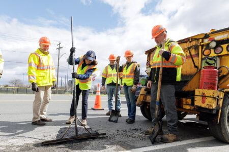 Road crew repairs asphalt with tools and a tar truck on a Finger Lakes road on a partly cloudy day.