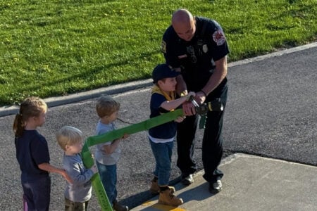 Firefighter guides three children holding a green fire hose on an asphalt surface.