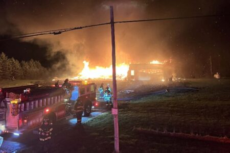 Firefighters battle a structure fire at night near a fire truck in a grassy field.