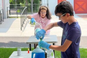 Two students wearing gloves pour liquids into containers during an outdoor science activity.