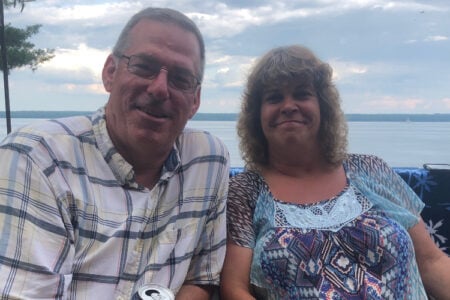 A man and woman smile outdoors with a lake in the background.