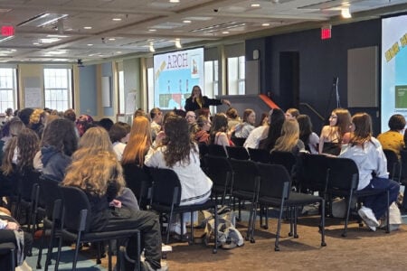A presenter speaks to an audience seated in chairs in a large room, with "ARCH" displayed on projection screens.