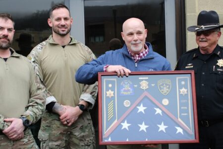 A man receives a framed display of patches and pins, plus a folded flag, from Steuben County Sheriff's Office personnel.