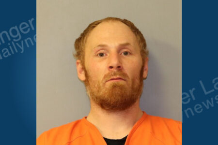 Mugshot of a man with red hair and a beard, wearing an orange jumpsuit against a gray background.