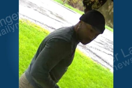 Dark-skinned man in black beanie and dark shirt bending over on grass near a paved surface, looking down.