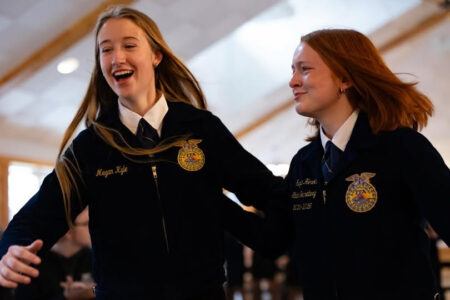 Two young women wearing navy blue FFA jackets with emblems, one laughing, at an indoor event.