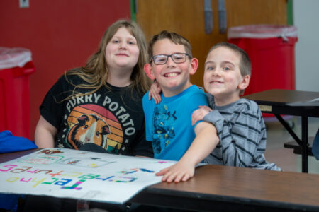 Three smiling students pose with their "Tech-tures" poster at a table in a school classroom.