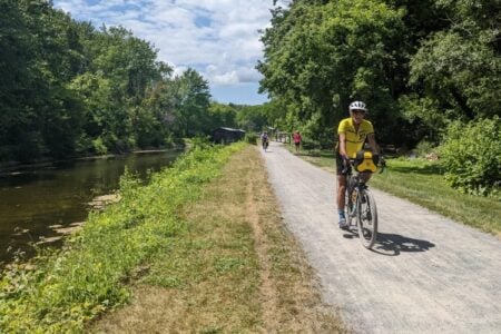 A person cycles on a scenic gravel trail alongside a canal, surrounded by lush green trees on a sunny day.