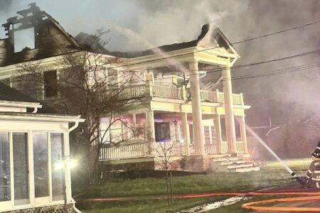 Firefighters spray water on a large, two-story house with roof damage and smoke, on a lawn at night.