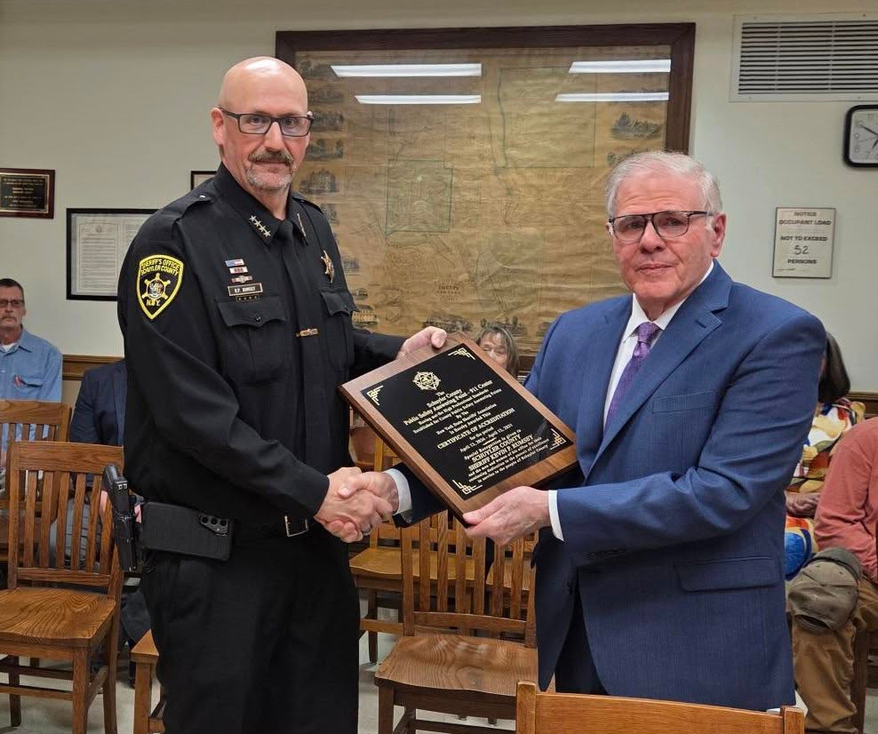 Schuyler County Sheriff Rumsey shakes hands with a man in a suit while receiving an accreditation plaque in a wood-paneled room.