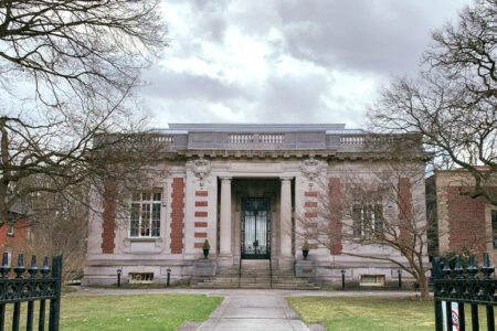 The Case Memorial Library, a stone building with brick accents and a wrought-iron gate, stands on a lawn under a cloudy sky.
