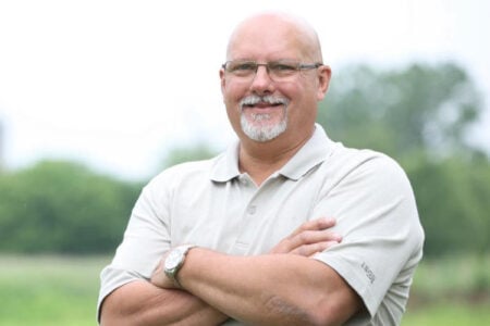 Headshot of a smiling man with glasses and goatee, arms crossed, outdoors.