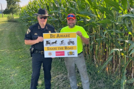 Sheriff and farmer pose with "Share the Road" sign in a rural area with cornfields for safety awareness.