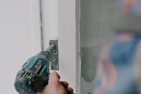 A hand uses a power drill to install a metal bracket into a white doorframe.