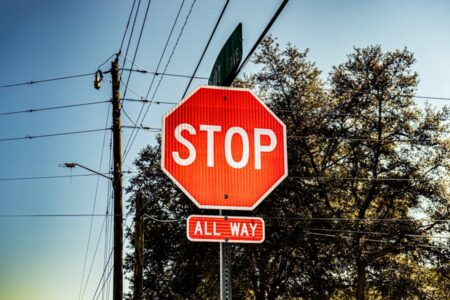 Red octagonal STOP sign with a smaller 'ALL WAY' sign below it, mounted on a pole against blue sky and trees.