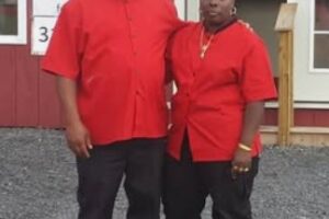 Black man and woman in matching red uniforms and headwear stand arm-in-arm outside a red business building.
