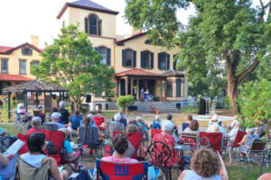 Audience watches outdoor theatrical performance on the porch of historic building in the Finger Lakes.