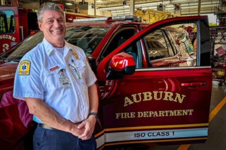 Smiling firefighter/EMT stands next to a red Auburn Fire Department vehicle inside a fire station.