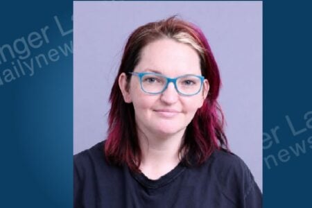 A woman with magenta hair and blue-rimmed glasses looks directly at the camera in a police-style portrait.