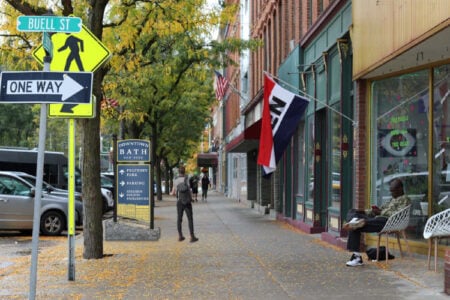 Autumn leaves line Buell Street in downtown Bath, NY, as a man walks and another sits outside a storefront.