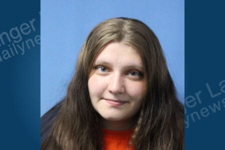 Mugshot of a young woman with long brown hair, blue eyes, an orange shirt, and a slight smile against a blue background.