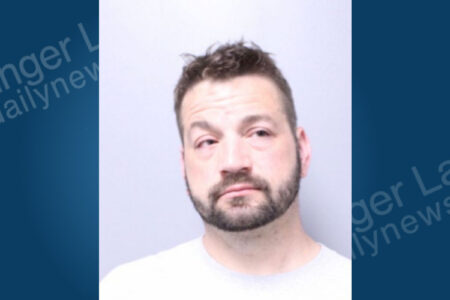 A man with dark, messy hair and a beard poses for a mugshot against a plain grey background.