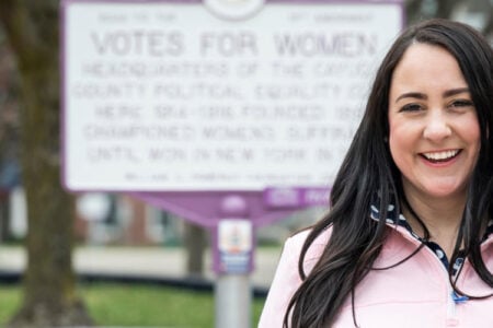 Smiling woman in a pink top poses outdoors near a historical marker commemorating "Votes for Women.