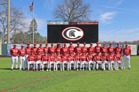 Cayuga CC Spartans baseball team in red uniforms poses on their home field with a logo scoreboard in the background.