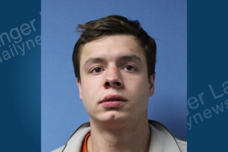 Young man with short brown hair and a neutral expression, wearing an orange and grey collared top, facing forward on a blue background.
