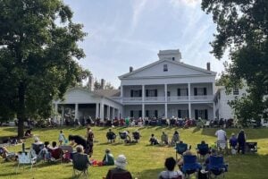 Crowd gathered on a sunny lawn in front of a grand white mansion with columns for an outdoor event.